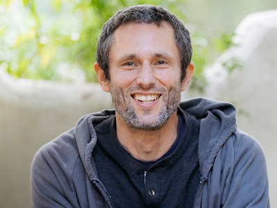 Charles Eisenstein smiling at the camera, and wearing a grey jacket in front of a lush green background. Charles Eisenstein is a writer and speaker focusing on the history of human civilization, consciousness, economics, ecology, and human cultural evolution.