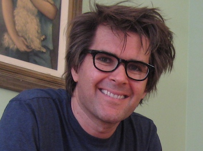 Mark Frauenfelder smiling in front of a green background with black glasses